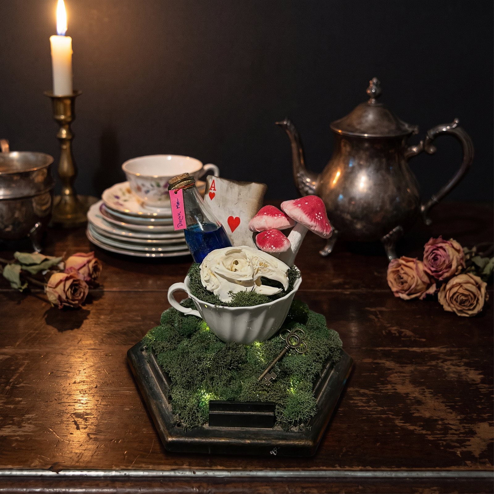 A small skull in a white teacup on lit moss, with red-capped mushrooms, a blue Drink Me bottle, a playing card and a brass key, set on a dark tea table by candlelight.