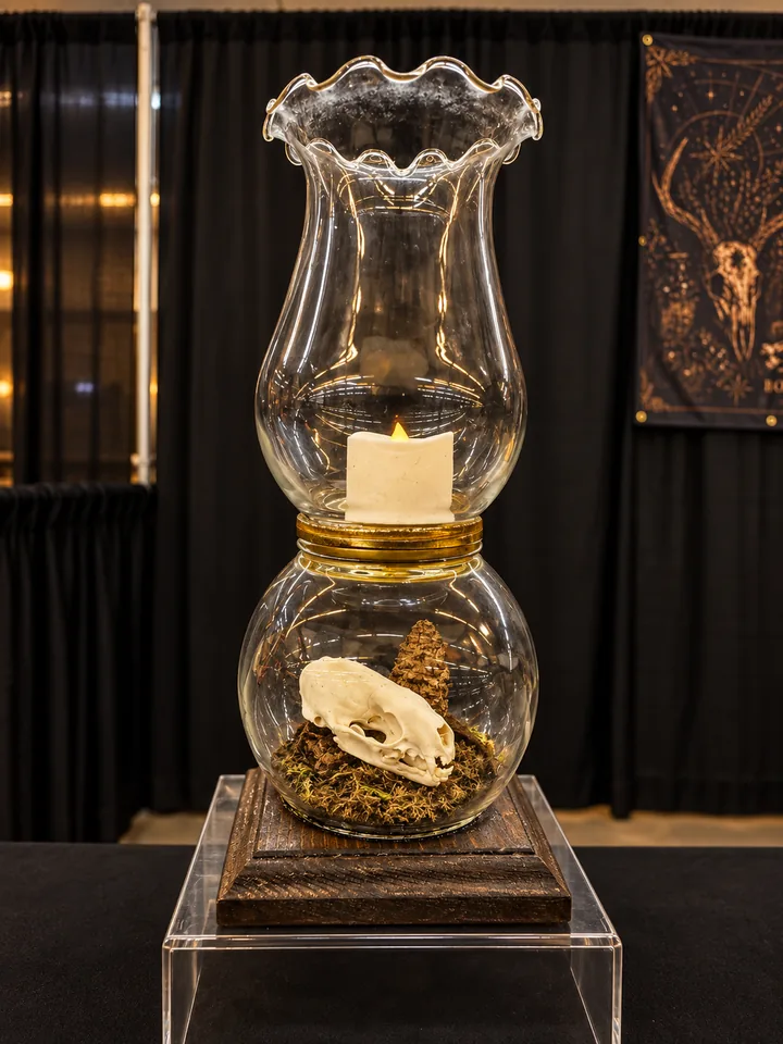 A glass hurricane lamp with a fluted rim: a flameless candle in the upper chamber and a small skull with a pine cone on moss in the round chamber below, on a dark wooden base.